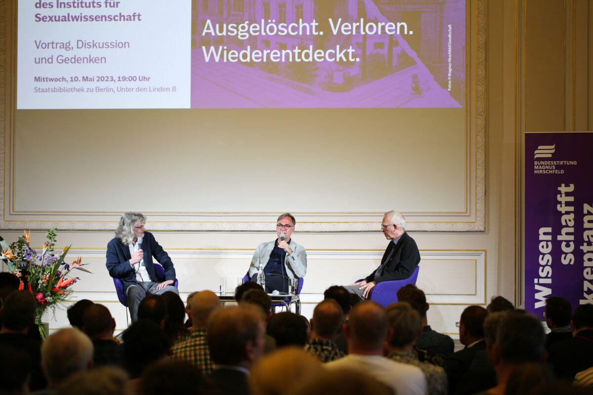 Foto. Drei sitzende Personen auf einer Bühne halten Mikrofone in der Hand und diskutieren miteinander. Am unteren Bildrand sind verschwommen viele Hinterköpfe zu erkennen.