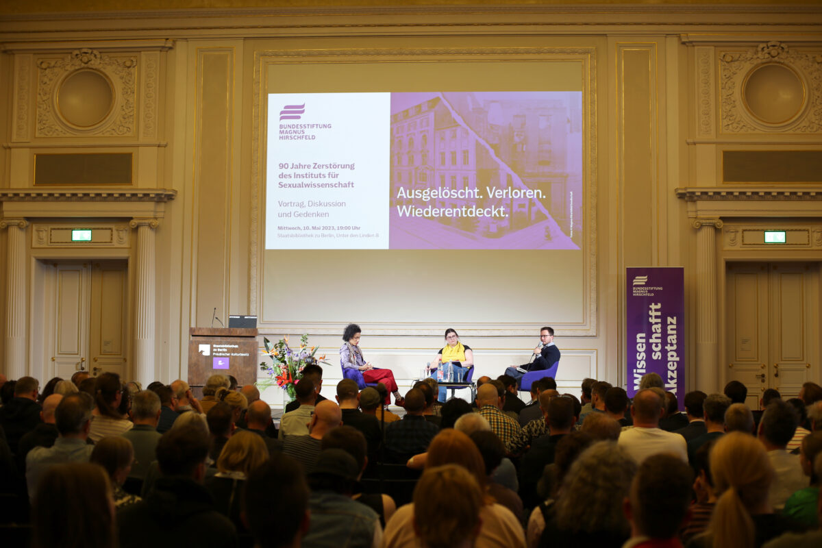 Foto. Drei sitzende Personen auf einer Bühne halten Mikrofone in der Hand und diskutieren miteinander. Am unteren Bildrand sind verschwommen viele Hinterköpfe zu erkennen.