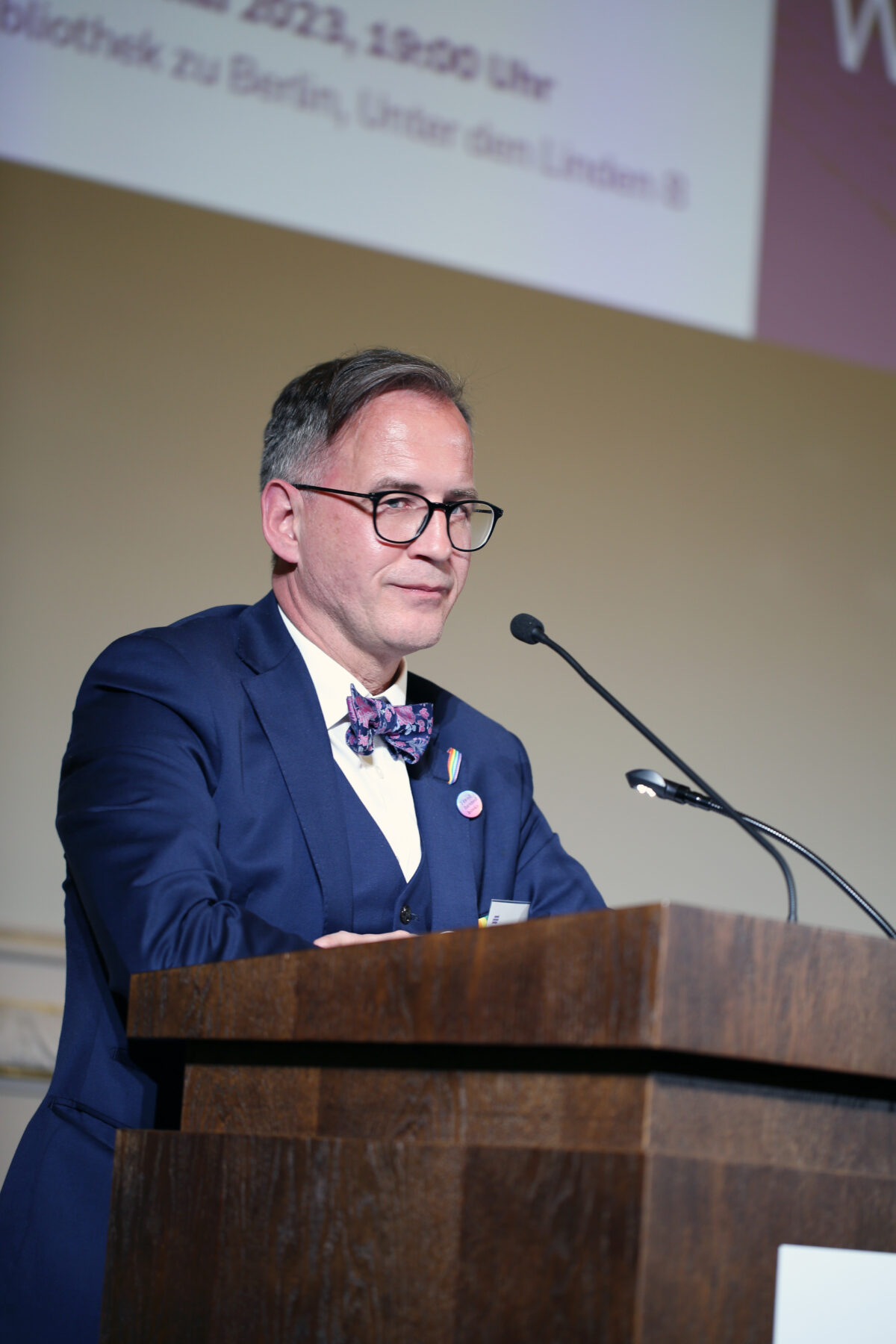 Foto. Eine Person (Helmut Metzner) redet von einer Bühne hinter einem Redner_innenpult. Gestaltungselement: Logo der Bundesstiftung Magnus Hirschfeld.