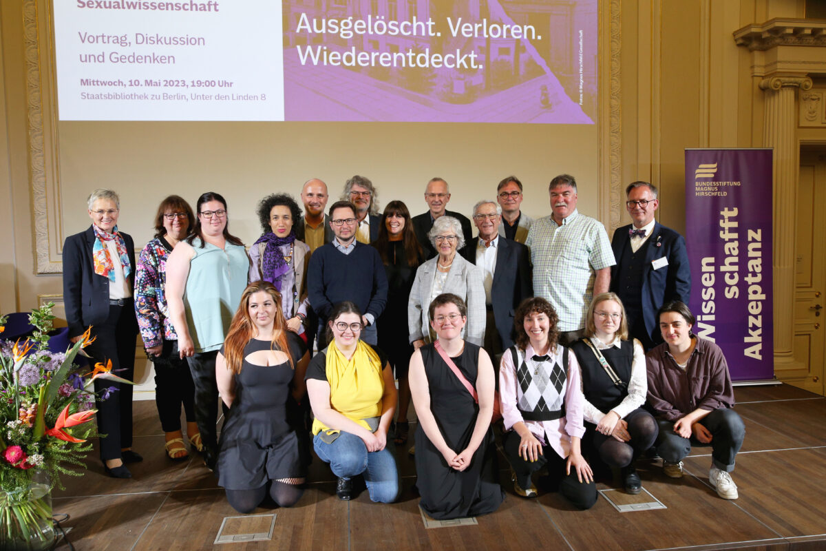 Foto. Auf einer Bühne stehen und sitzen in insgesamt drei Reihen 20 Personen und lächeln in die Kamera. Am linken Rand ist ein großer Blumenstrauß zu sehen. Am rechten Bildrand steht im Hintergrund ein hochformatiger Aufsteller mit der Aufschrift