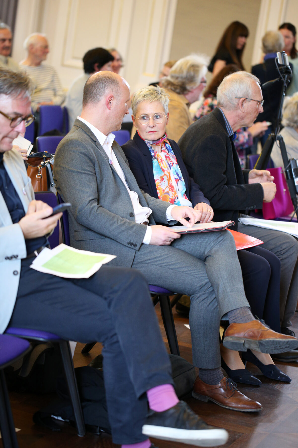 Foto. Zwei Personen (Sven Lehmann, Ruth Schröder) sitzen in einer Stuhlreihe, haben die Köpfe zu einander geneigt und unterhalten sich. Links und rechts sind unscharf weitere sitzende und stehende Personen zu erkennen.