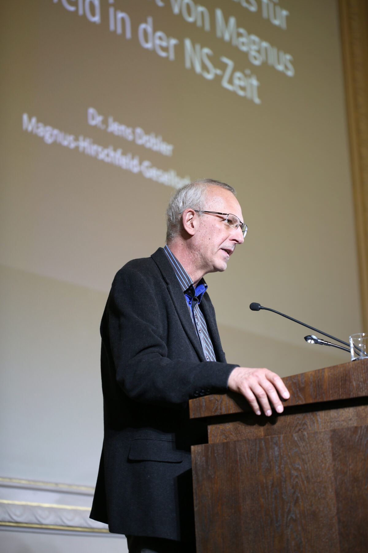 Foto. Eine Person steht hinter einem Redner_innenpult und spricht von einer Bühne. Im Hintergrund ist eine schwarz-weiße Textprojektion an der Rückwand der Bühne zu erkennen. Der Text ist zum Teil unscharf. Zu lesen sind noch die Worte „NS-Zeit“ und „Magnus“.