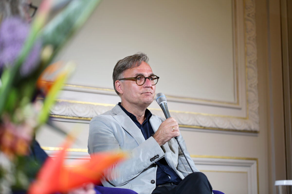 Foto. Eine Person sitzt und hält ein Mikrofon in der Hand. Am linken Bildrand ist im Anschnitt unscharf ein Blumenstrauß im Vordergrund zu sehen.