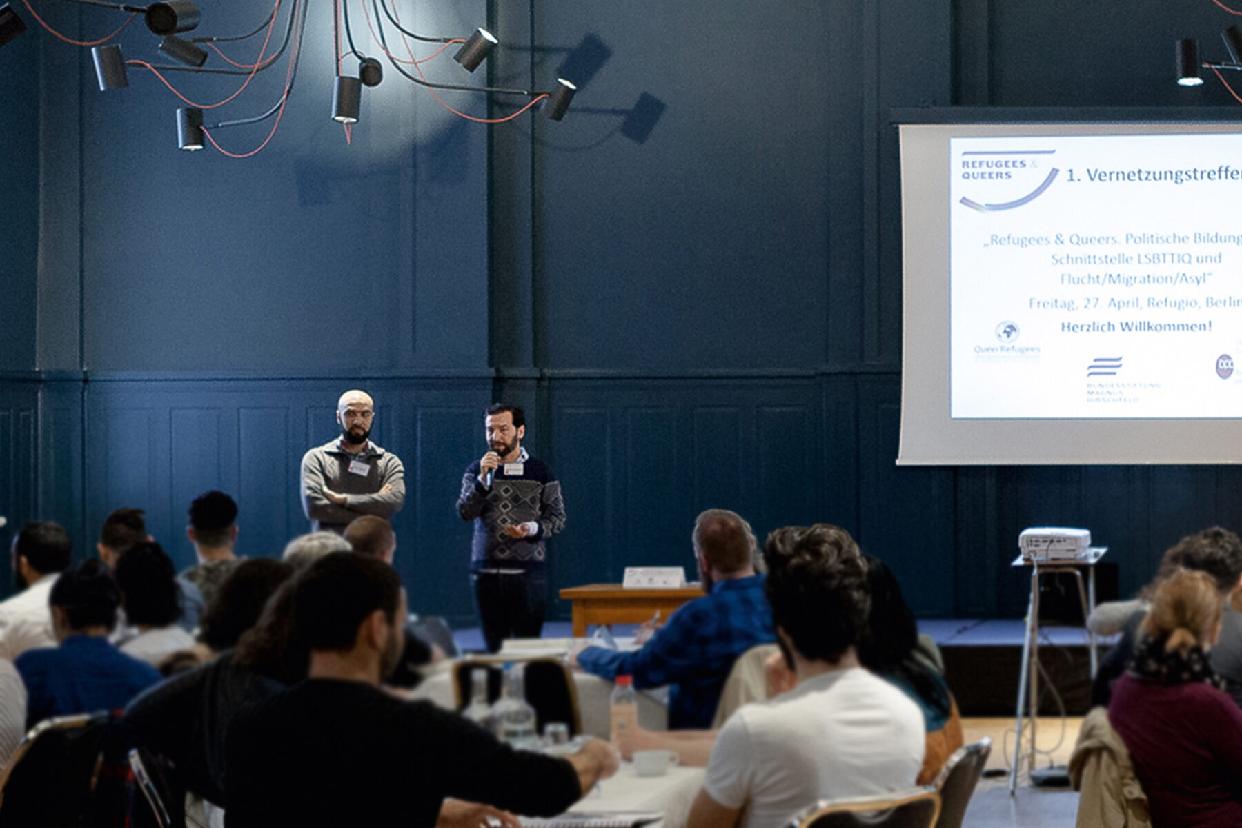 Foto: eines gefüllten Vortragssaals. Im Vordergrund befinden sich verschwommen zuhörende Personen. Die Personen sitzen an Tischen. Im Hintergrund stehen zwei Personen. Die rechte Person spricht in ein Mikrofon. Rechts am Bildrand ist ein Beamer, eine Leinwand und eine laufende Präsentation.