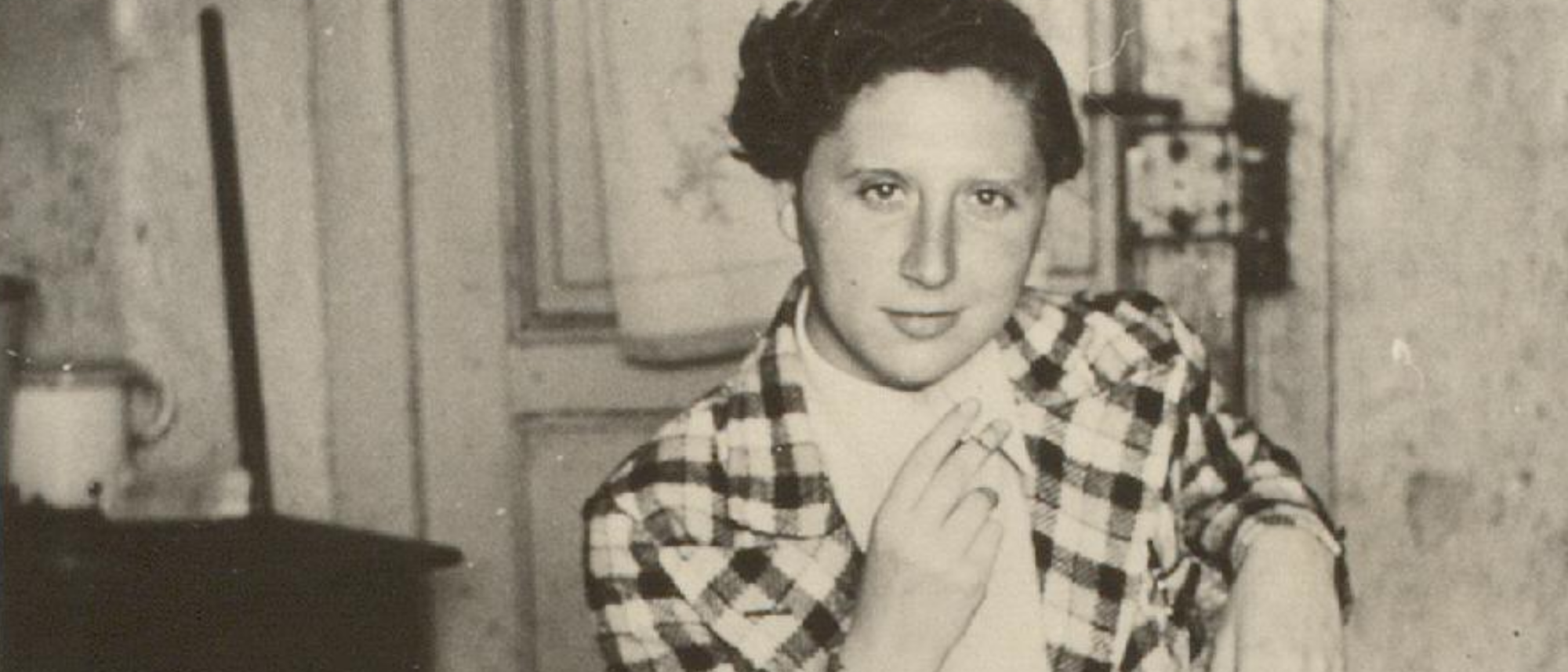 Schwarz-weiß Foto einer Person, die auf einem Holzstuhl sitzt und in die Kamera schaut. Die Person hat ihren rechten Arm leicht auf dem Oberschenkel aufgestützt. Im Hintergrund ist ein Schrank und eine Zimmertür zu erkennen.