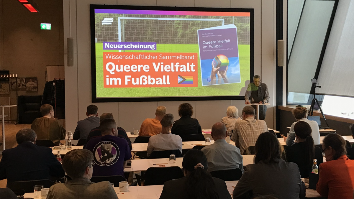 Foto von Menschen in Seminarraum. Neben einer großen Bildprojektion steht eine Person, die zum Publikum spricht. Im Vordergrund sitzen zuhörende Personen, die nur von hinten zu sehen sind. Die Projektion zeigt eine Textgrafik. Darauf ist vor einem Fußballtor das Cover des Sammelbands 