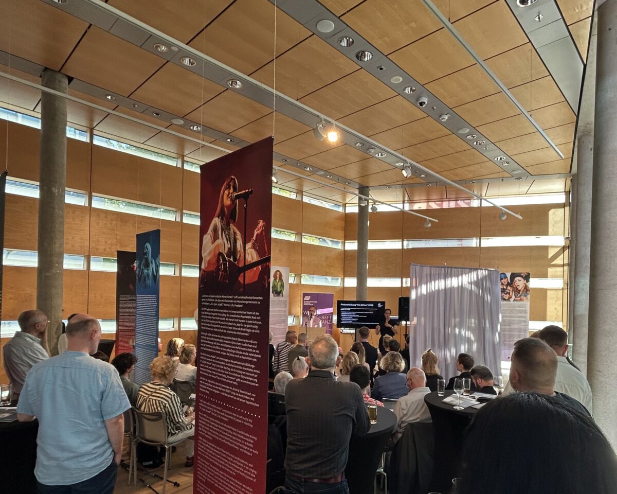 Foto. Zu sehen ist: Draufsicht auf die Verleihung des Iris-Orfeo-Award in der Schwedischen Botschaft Berlin. Am Mikrofon BMH-Bundesvorstand Helmut Metzner. Im Vordergrund: sitzendes Publikum.