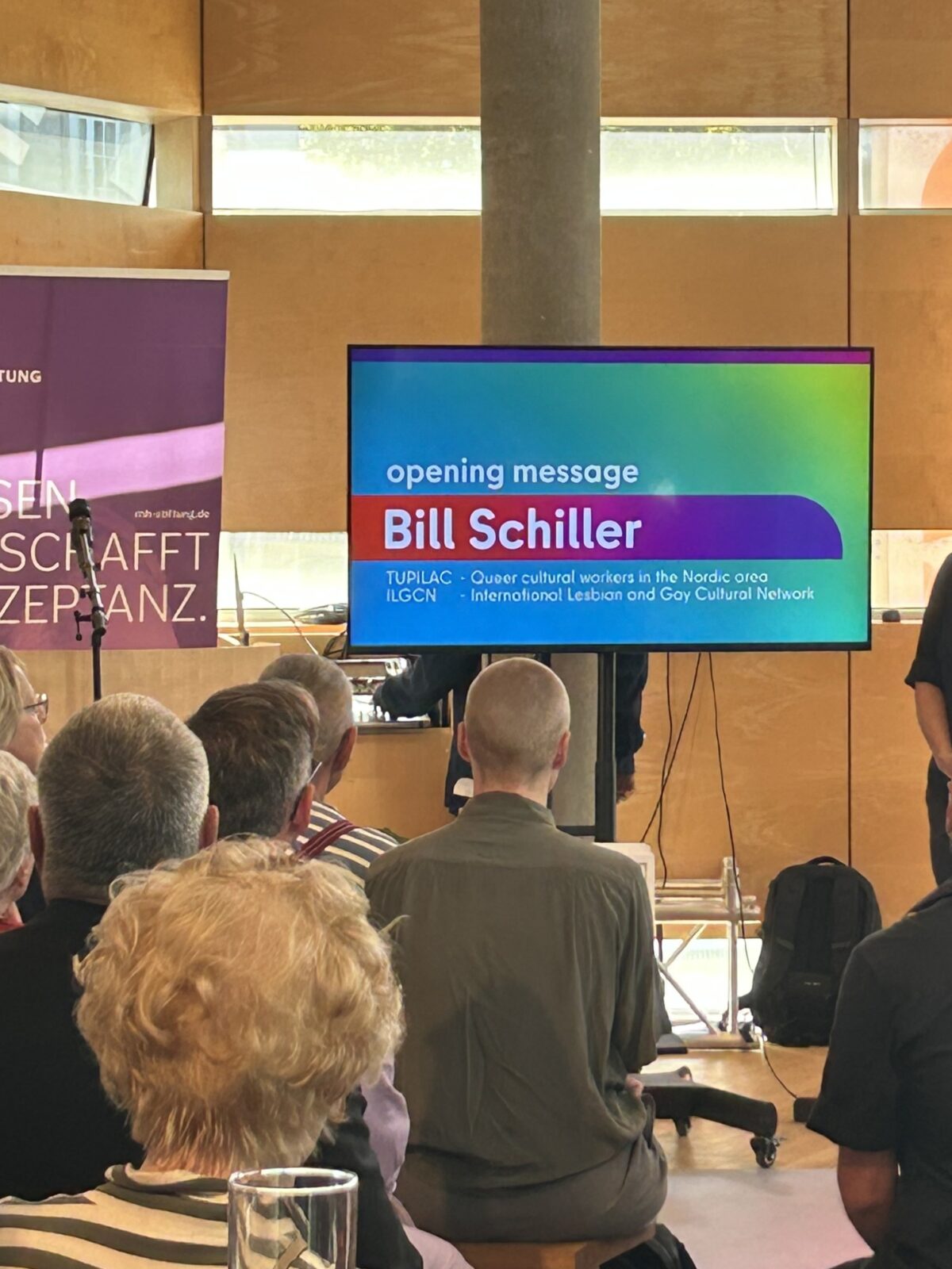 Foto. Zu sehen ist ein Monitor mit der Eröffnungsrede von Bill Schiller. Im Vordergrund: sitzendes Publikum.