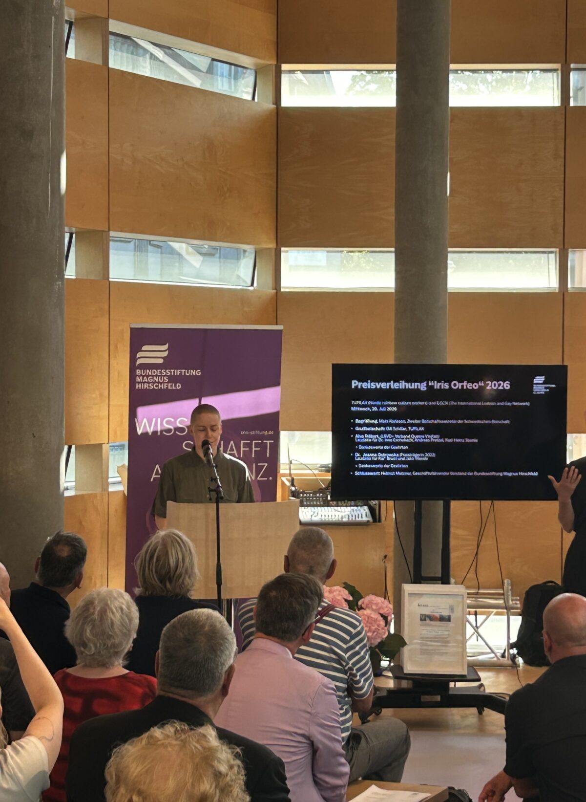 Foto. Zu sehen ist Laudatorin Alva Träbert am Mikrofon im Rahmen der Iris-Orfeo-Award-Preisverleihung in der Schwedischen Botschaft Berlin. Im Vordergrund: sitzendes Publikum.