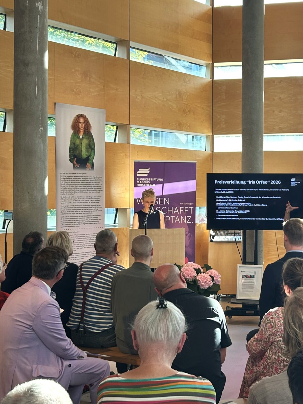 Foto. Zu sehen ist: eine Person am Mikrofon im Rahmen der Verleihung des Iris-Orfeo-Award in der Schwedischen Botschaft Berlin. Im Vordergrund: sitzendes Publikum.