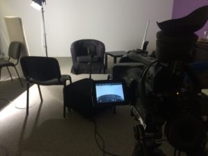 Foto: von einer Interview-Aufnahme. Es zeigt einen Raum mit mehreren Stühlen und Technik. Im Vordergrund ist ein Kamerastativ. Die Kamera befindet sich im Aufnahmemodus. Ein links im Bild stehender Scheinwerfer erleuchtet Teile des Raums.