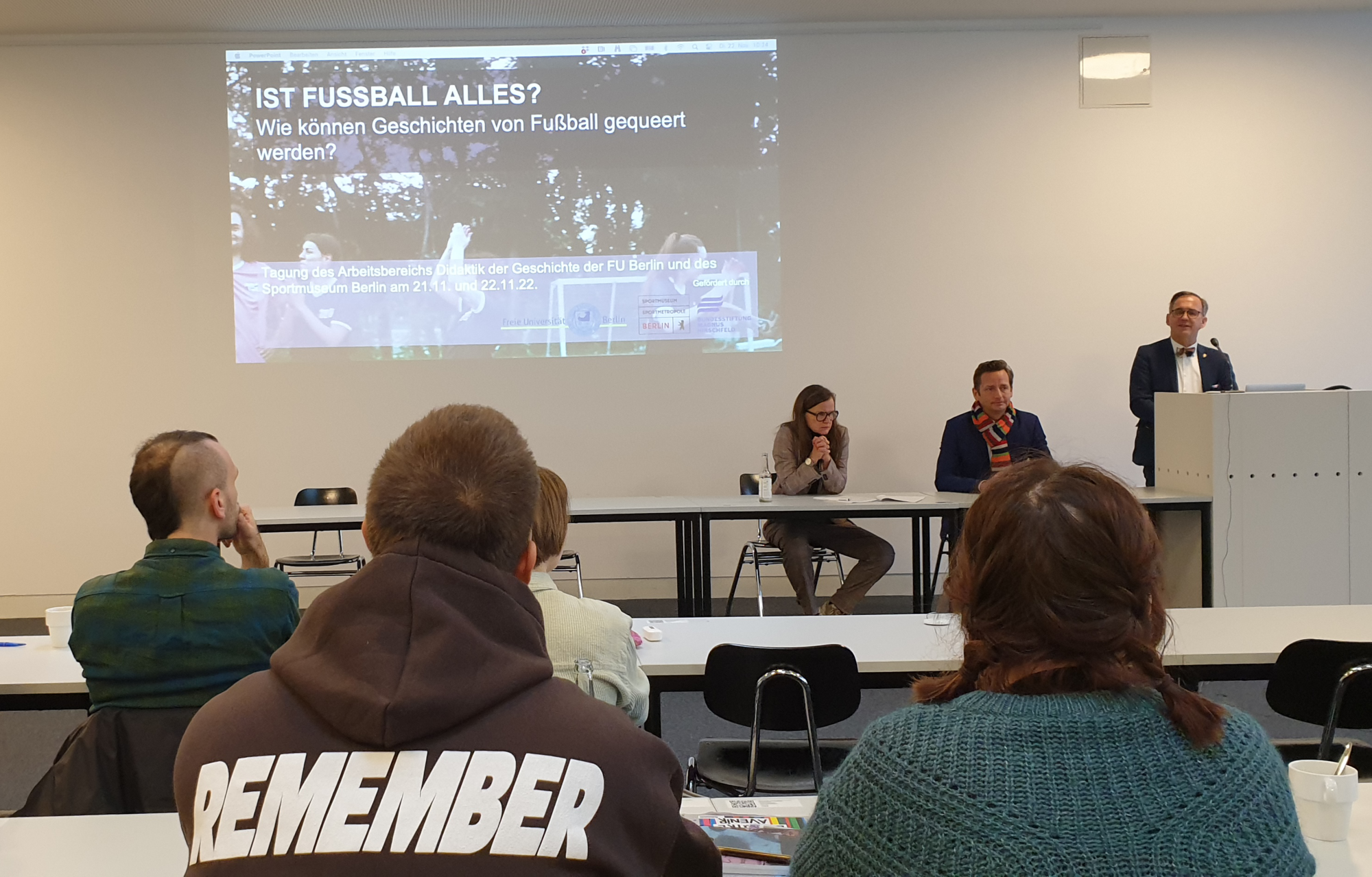 Foto von Menschen in Seminarraum. Auf dem Podium sitzen 2 Personen und 1 Person hält einen Vortrag. Im Vordergrund sitzen einige zuhörende Personen, die nur von hinten zu sehen sind.
