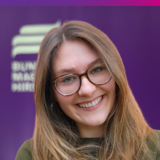 Portraitfoto Carolin Beinroth Im Hintergrund ist verschwommen das Logo der Bundesstiftung Magnus Hirschfeld in weiß vor lila Hintergrund zu erkennen.